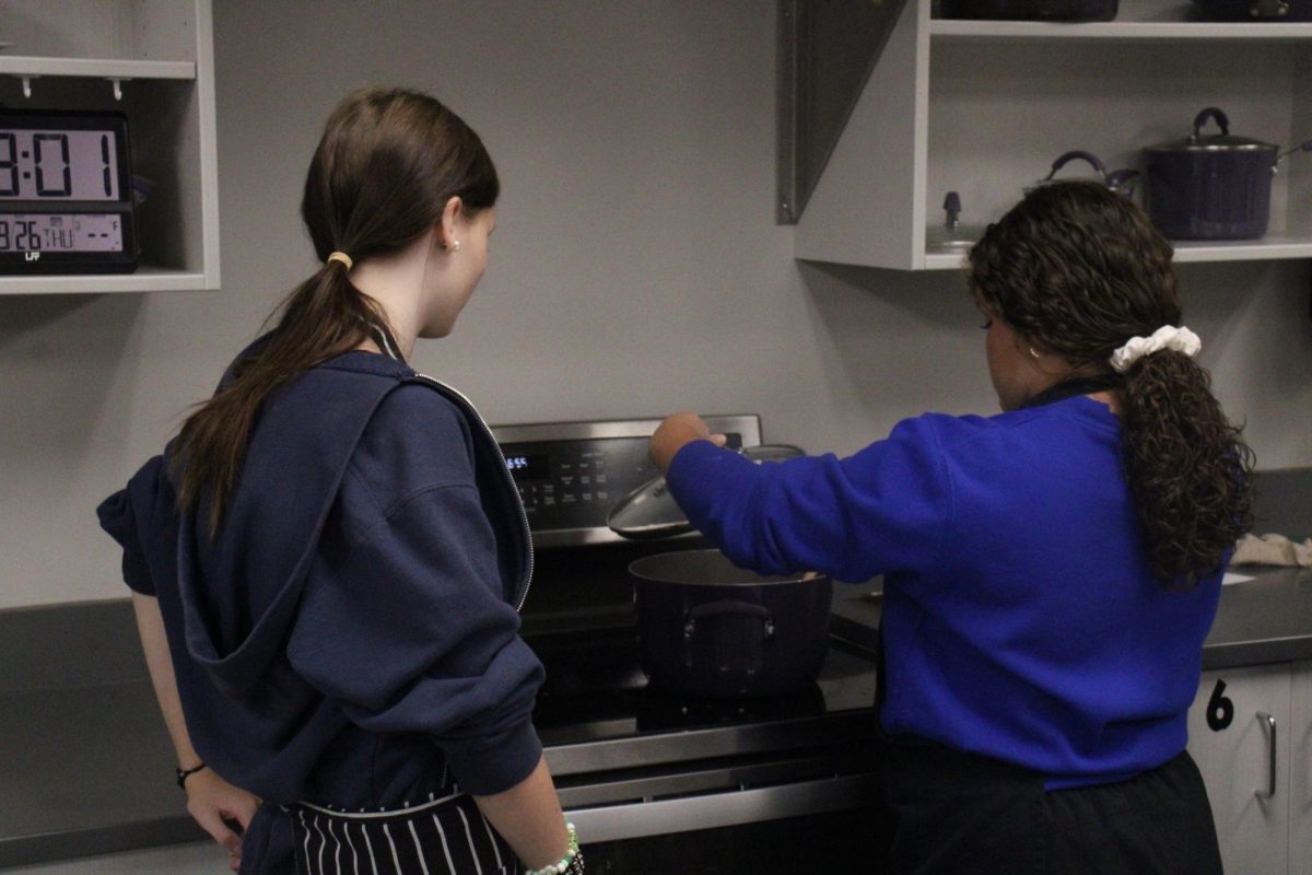 A student checks on something on the stove while another student stands by.