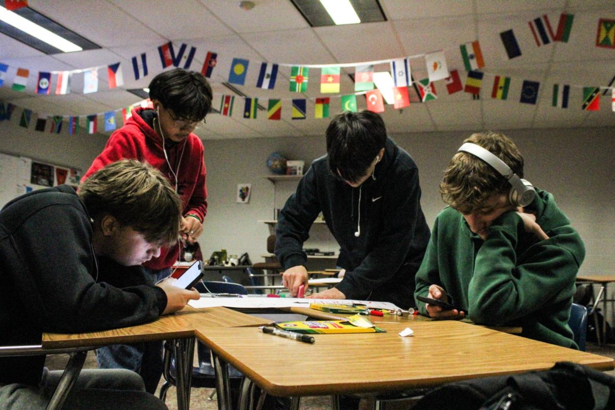 Two students work on poster while two play on their phones. 