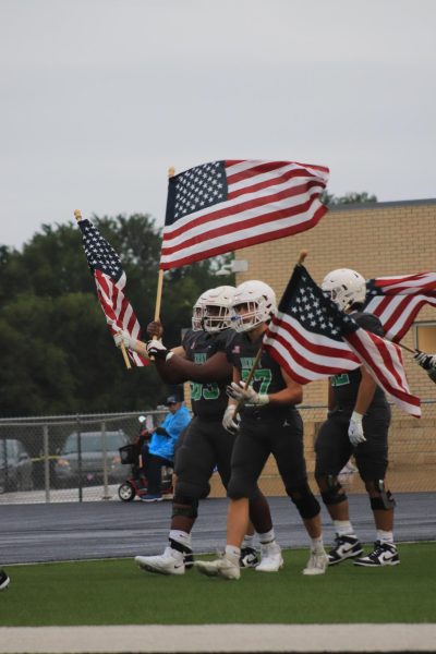 Football players walk onto field holding flags.