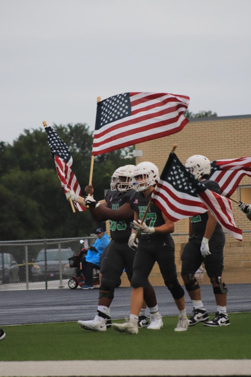 Football players walk onto field holding flags.
