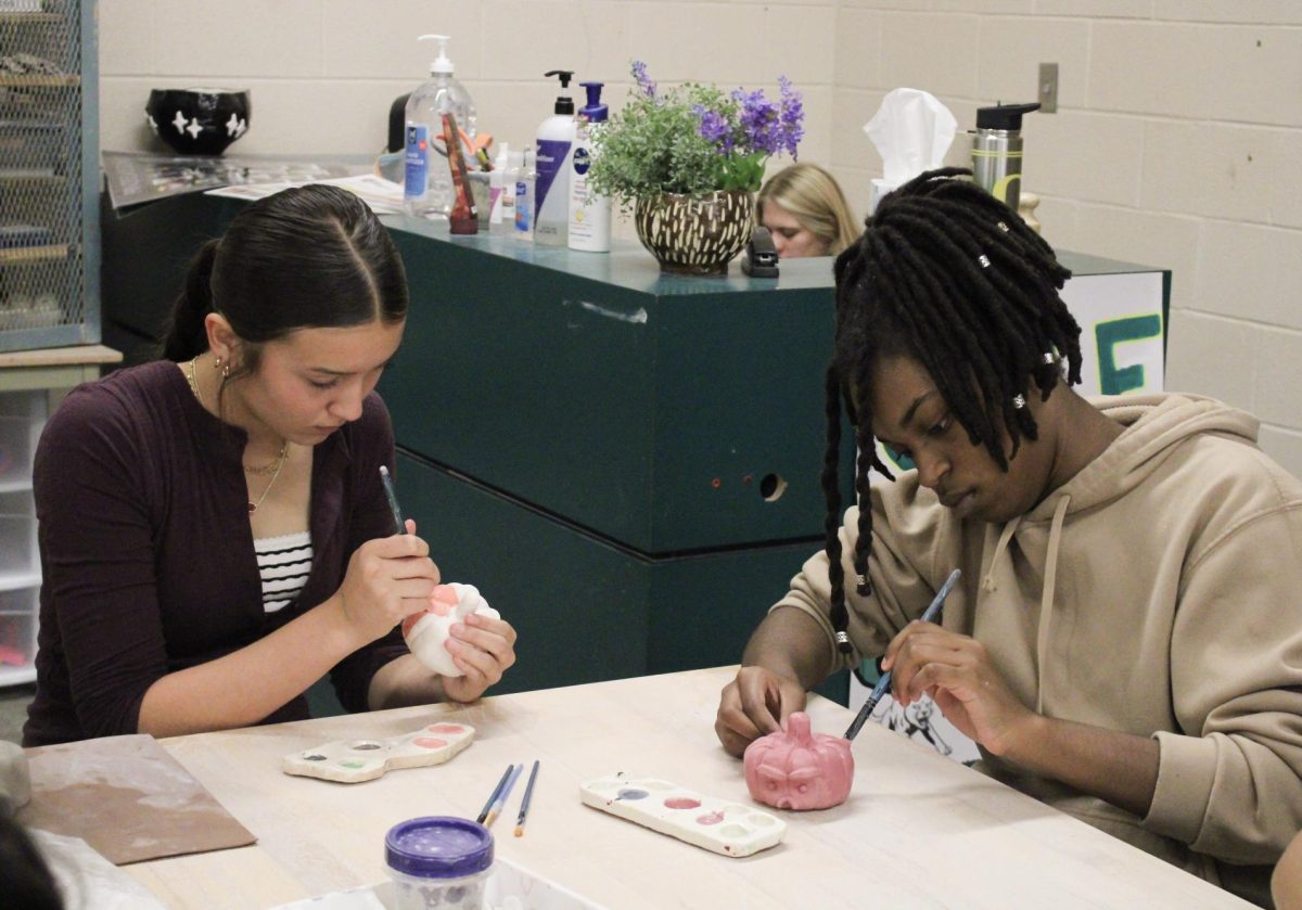 Two students are seen painting over their pumpkins with a pink paint for their projects in Clay Fundamentals.